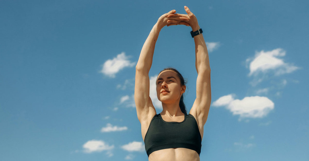 Woman stretching outdoors under a blue sky with white clouds. Her arms are raised above her head, showing her athletic build and fitness apparel. This image represents biohacking for women's health and wellness through exercise and mindfulness.