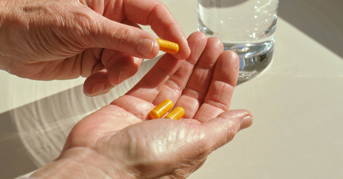 A close-up of a person's hand holding three yellow dietary supplement capsules with a glass of water in the background. Shows someone taking their daily vitamins or supplements.