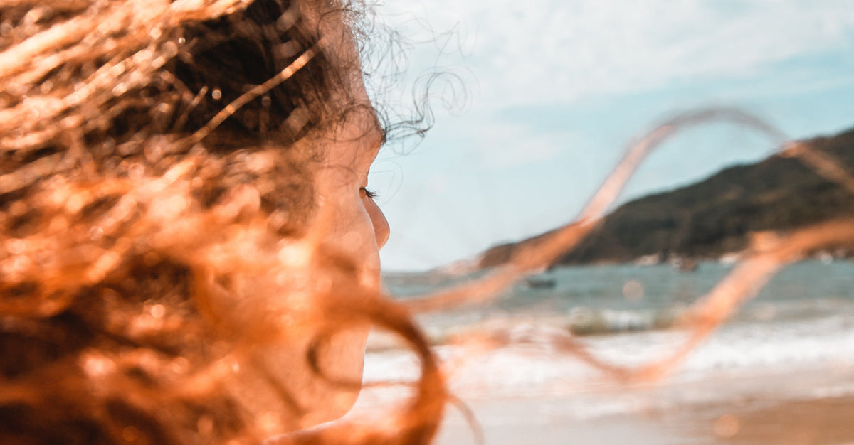 A profile view of a person with curly hair blowing in the wind, looking out at a beach and the ocean.