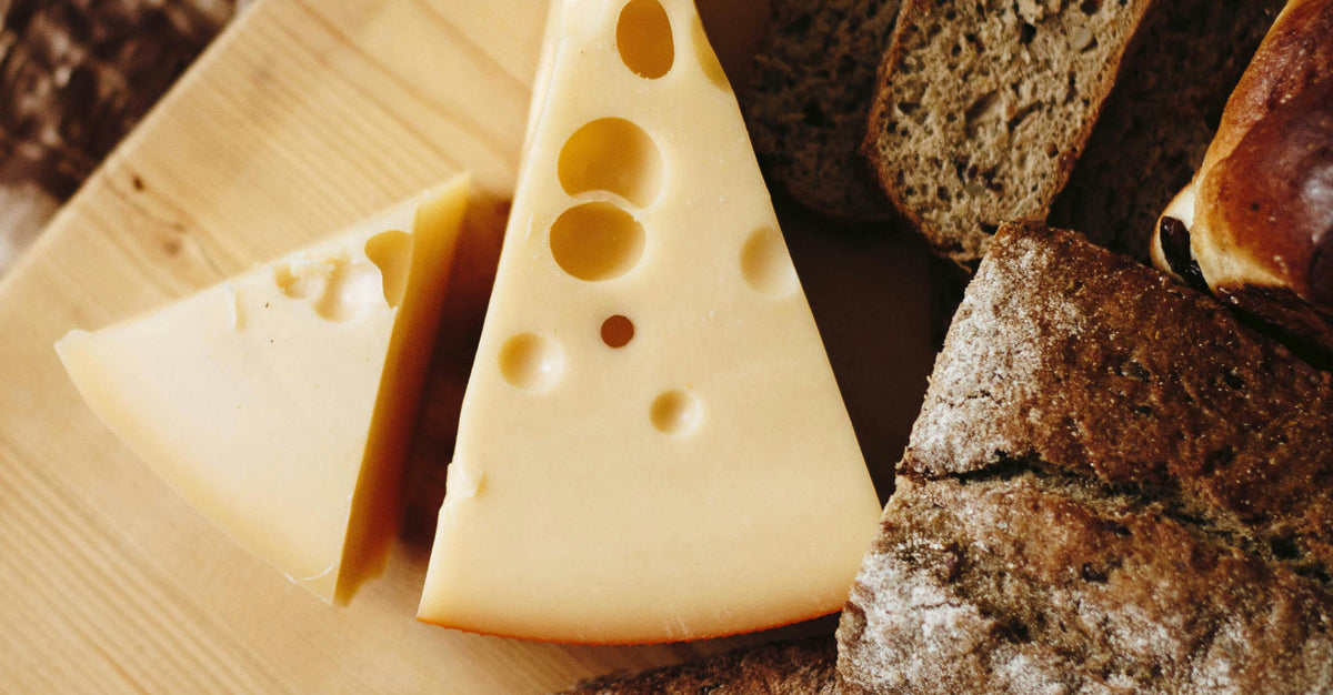 A close-up, overhead shot of a triangular wedge of Swiss cheese, or a similar aged cheese with holes, next to two slices of dark, whole-grain bread on a wooden cutting board.