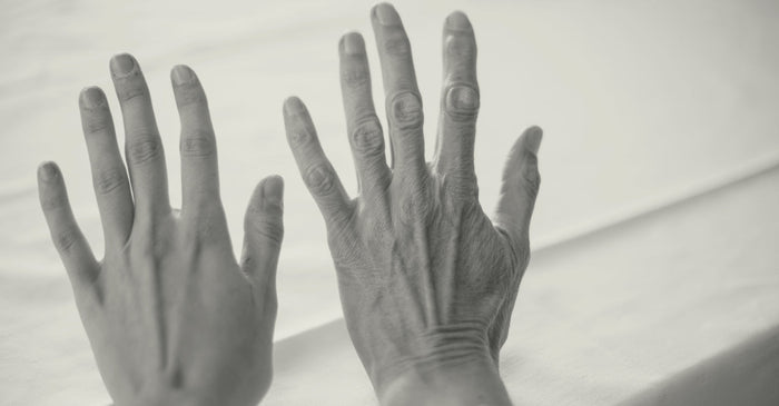 A black and white photo of two hands, one young and one old, placed side-by-side on a light-colored surface. The young hand is on the left, and the older, wrinkled hand is on the right, illustrating the physical effects of aging.