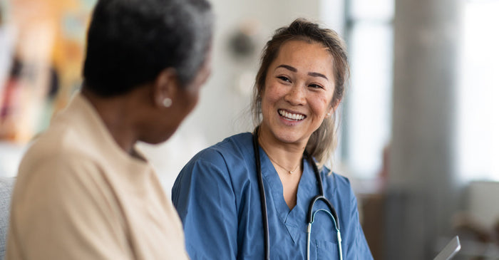 A smiling female healthcare professional in blue scrubs talks with an older female patient.
