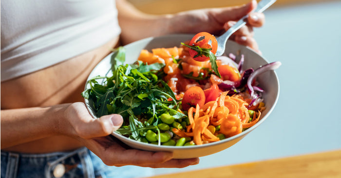 A person holding a colorful bowl of fresh vegetables, including arugula, cherry tomatoes, shredded carrots, edamame, and salmon, with a fork lifting a bite.