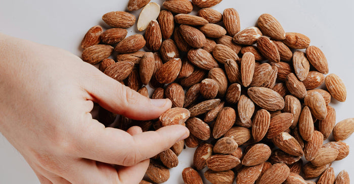 A hand reaching into a large pile of almonds, picking one up with the thumb and forefinger.