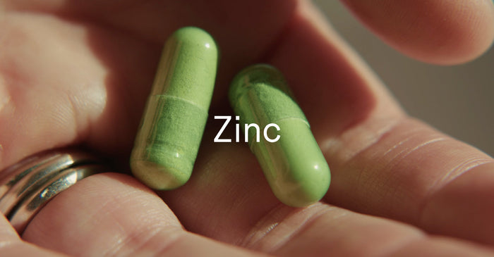Close-up of two green zinc capsules resting on a person’s palm with the word “Zinc” overlaid in white text.