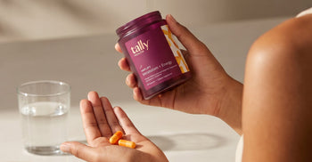 A person holding a jar of Tally Health Amplify Metabolism + Energy supplement while tipping orange capsules into their palm, with a glass of water nearby.