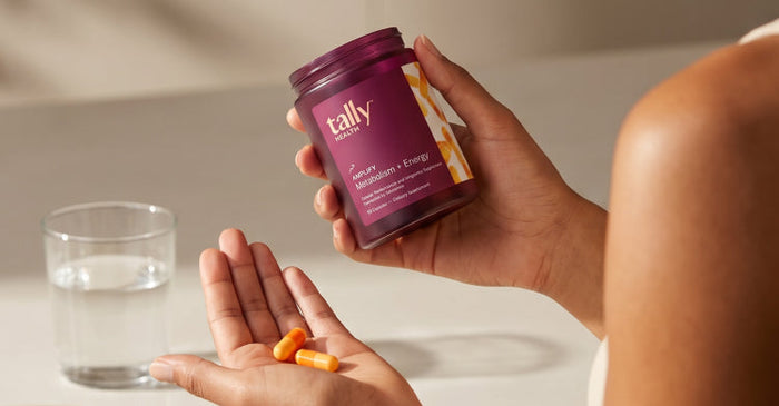 A person holding a jar of Tally Health Amplify Metabolism + Energy supplement while tipping orange capsules into their palm, with a glass of water nearby.
