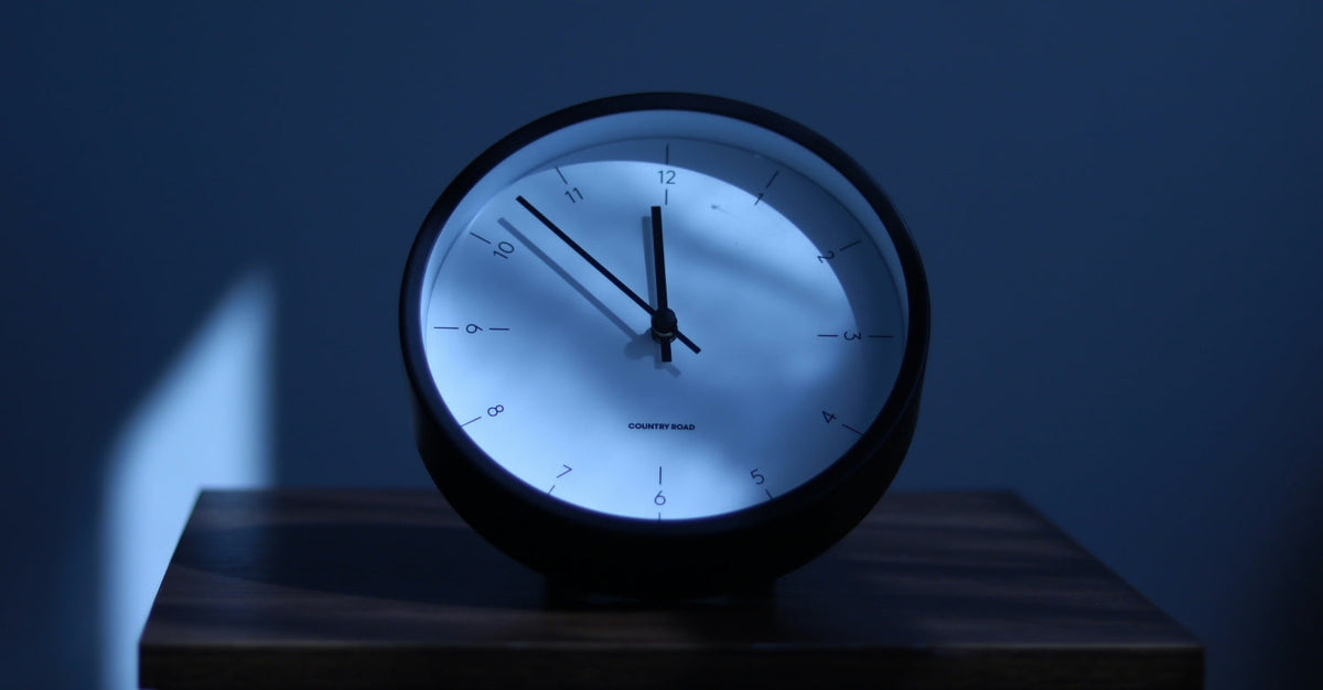 A round black alarm clock glowing with blue-white light on a wooden nightstand at night, showing the time as approximately 12:37