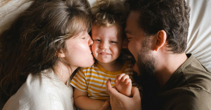 An overhead photo of a mother and father in bed, both kissing their smiling toddler on the cheek.