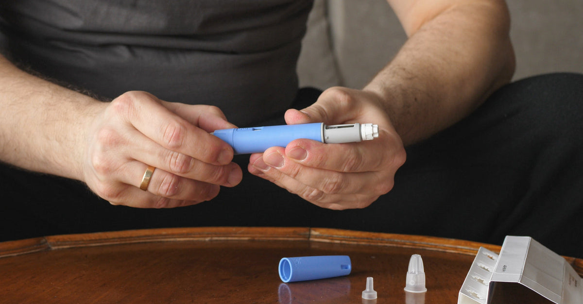 A close-up shot of a person's hands holding a GLP-1 injection pen. This image features a person's hands manipulating a blue and white injector pen. The pen is being assembled or prepared for use, with the cap and needle cover visible on the table. The ima