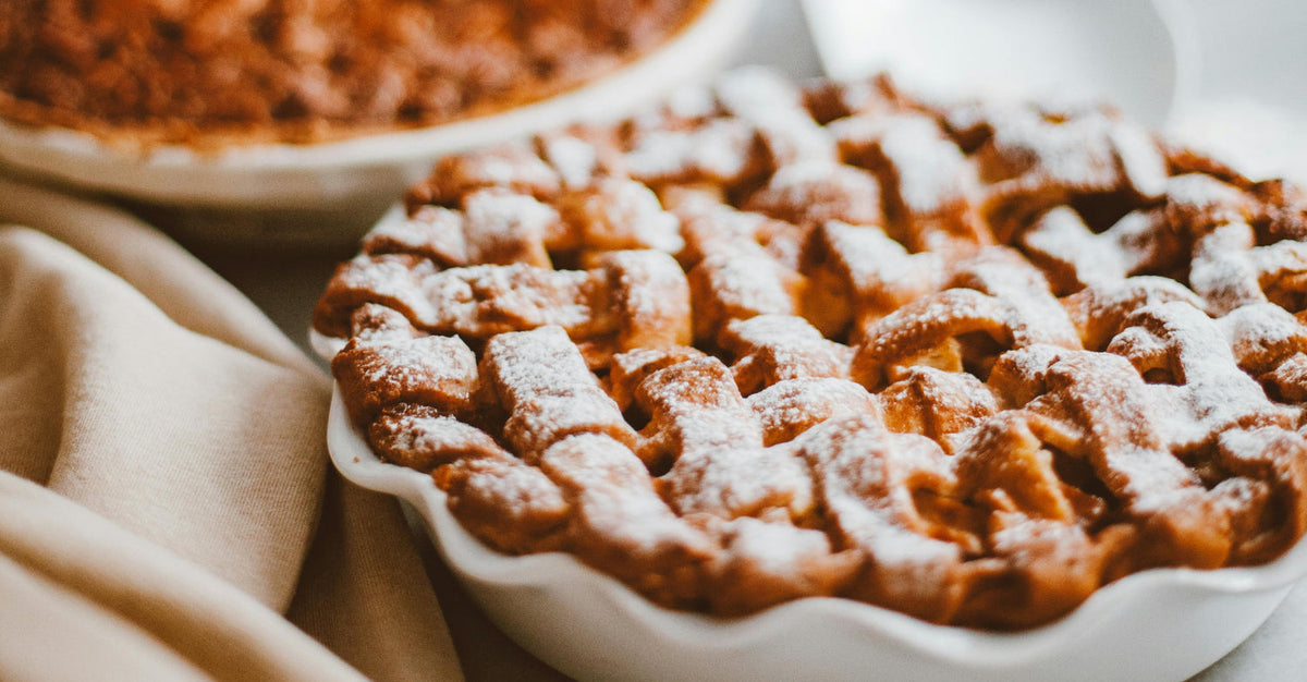 Close-up of a golden-brown lattice-topped pie dusted with powdered sugar, with another pie blurred in the background on a cozy beige cloth.