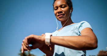 A woman outdoors checking her smartwatch while wearing earbuds, with a clear blue sky in the background.