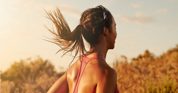 A woman with a ponytail, wearing a pink running top, is shown from the side as she runs outdoors with her back to the camera. The setting sun shines on her.
