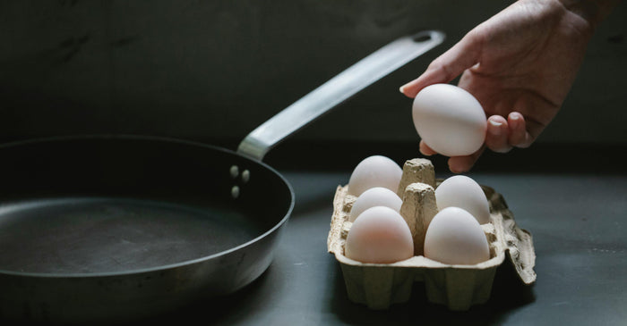 A person's hand is shown turning a knob on a black gas stove with a nonstick frying pan on the burner. The pan is on the heat and appears to have a small amount of oil in it.