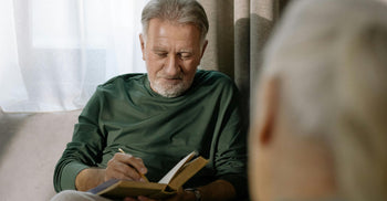 Older man sitting indoors and writing in a notebook, symbolizing reflection, cognitive health, and proactive lifestyle choices for healthy aging.