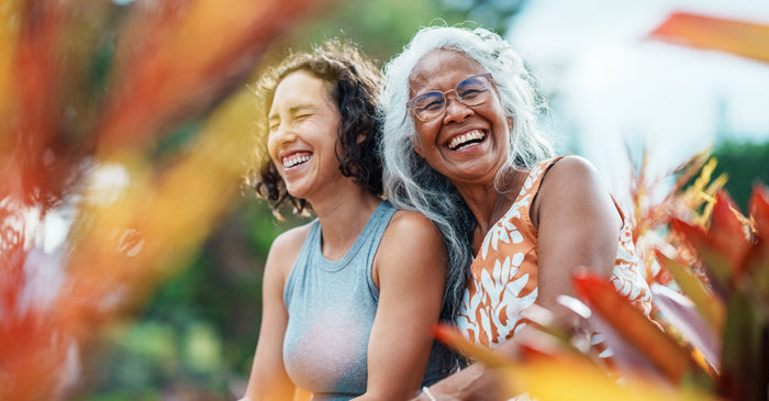 Older and younger women sitting together outdoors laughing, symbolizing intergenerational wellness, resilience, and long-term healthspan.
