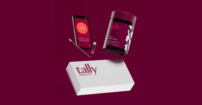 A still-life photo against a dark burgundy background. A Tally Health kit box, a floating supplement bottle, and a smartphone are all arranged in the center. The smartphone screen displays the Tally Health app, showing a 