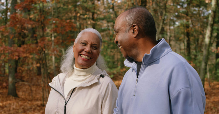 An older couple walks outdoors in a forest with fall foliage. Both are smiling warmly at each other, wearing light jackets and enjoying a peaceful moment together.