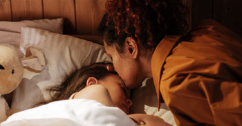 Mother kissing her sleeping child's forehead in bed at night, with a plush toy on the pillow beside them.