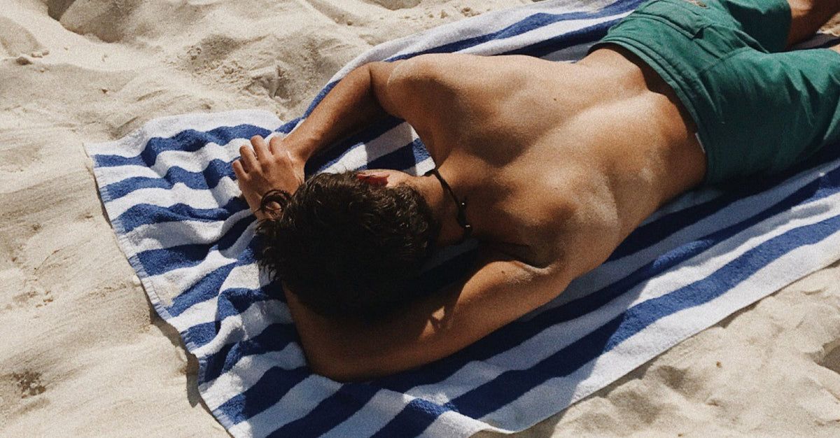 Man lying shirtless on a striped beach towel in direct sun, illustrating UV exposure as a summer stressor on the body.