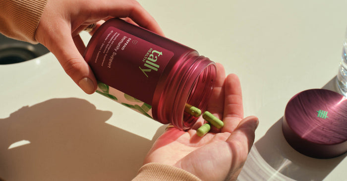 A person's hands are shown pouring green capsules from a dark red bottle labeled 