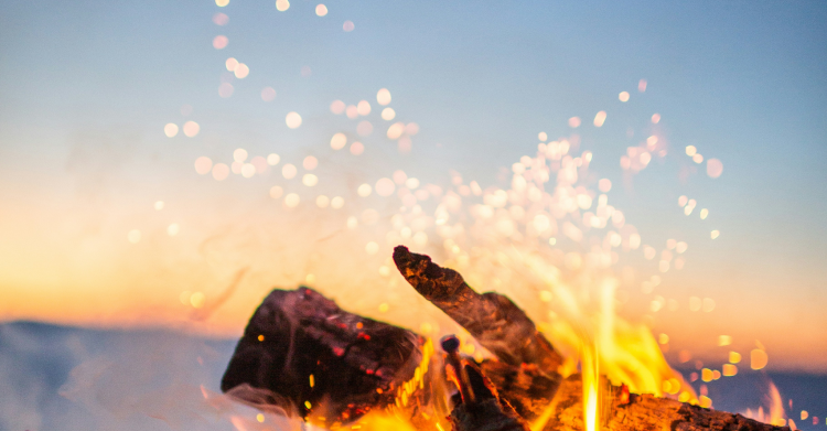 Close-up of a crackling campfire with glowing embers and sparks rising against a dusky sky.