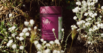 Tally Health DEFEND Immunity Support supplement canister nestled among small white wildflowers and green foliage outdoors