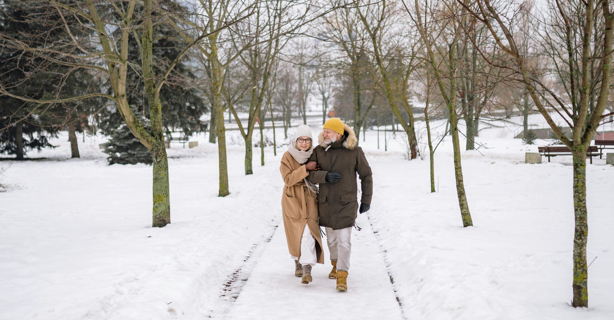 Older couple walking arm in arm through a snow-covered park, bundled in winter coats and hats.