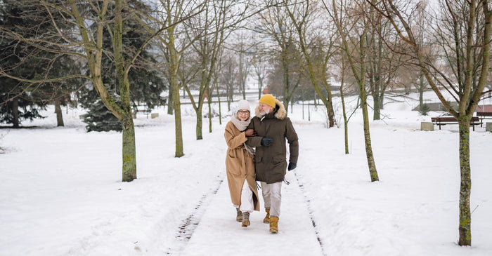Older couple walking arm in arm through a snow-covered park, bundled in winter coats and hats.