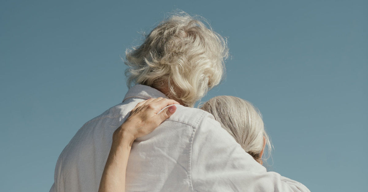 Two older adults with silver hair photographed from behind, one resting their head on the other's shoulder, set against a clear blue sky — evoking connection, longevity, and shared life.