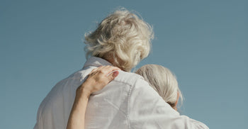 Two older adults with silver hair photographed from behind, one resting their head on the other's shoulder, set against a clear blue sky — evoking connection, longevity, and shared life.