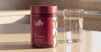 A maroon Tally Health NAD+ Support supplement bottle sitting on a sunlit countertop next to a glass of water.